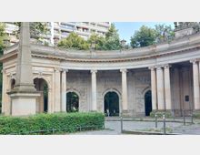 Halbrunde steinerne Kolonnaden mit verzierten Säulen und Reliefs, flankiert von einem Obelisken links im Vordergrund; im Hintergrund Bäume und moderne Wohngebäude sichtbar.