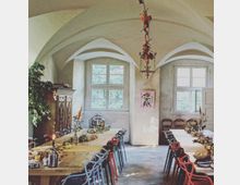 Ein historischer Raum mit Kreuzgewölbedecke und großen Sprossenfenstern, eingerichtet mit einer langen Tafel, die mit Dekorationen und Geschirr gedeckt ist. An den Tischen stehen bunte Stühle, und an der Decke hängt ein verzierter Kronleuchter; links im Raum steht ein antiker Holzschrank, rechts sind drei Stühle an der Wand aufgereiht.