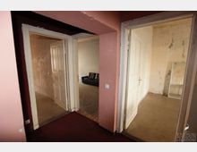 Flur in einem alten Gebäude mit drei offenen Türen, die in Räume mit unterschiedlichen Bodenbelägen führen; links eine Tür in einen Raum mit Betonboden, mittig ein Raum mit hellem Teppich und schwarzem Sofa, rechts ein Raum mit Spiegel an einer abgenutzten, hellen Wand.