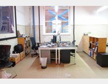 Arbeitsplatz in einem hellen Büroraum mit weiß gefliesten Wänden, einem großen Fenster und beige gefliestem Boden. Im Raum stehen ein Schreibtisch mit Bürostühlen, Regale mit Büchern und Unterlagen sowie diverse Büromaterialien.