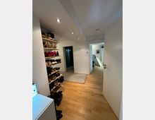 Flur in einer Wohnung mit hellem Holzfußboden, weißen Wänden und eingelassenen Deckenstrahlern. Auf der linken Seite befindet sich ein offenes Schuhregal mit mehreren Reihen von Schuhen, während rechts eine geöffnete Tür den Blick in einen anderen Raum freigibt.