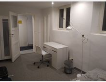 Ein schlicht eingerichtetes Zimmer mit einem Schreibtisch, einem weißen Drehstuhl und einer Stehlampe. Im Hintergrund sind ein Kleiderschrank, eine geöffnete Tür und zwei schmale Fenster an der weißen Wand zu sehen.