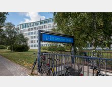 Eingang zur U-Bahn-Station „Amrumer Straße“ mit einem blauen Stationsschild, umgeben von grünen Bäumen und Fahrrädern, im Hintergrund ein mehrstöckiges Bürogebäude mit Glasfronten.