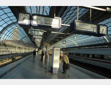 Ein moderner Bahnhof mit einem gewölbten Glasdach, das Tageslicht einlässt. Fahrgäste stehen auf den Bahnsteigen, ein Zug steht auf einem Gleis, und Anzeigetafeln sowie Uhren zeigen Informationen zu Abfahrten und Gleisen.