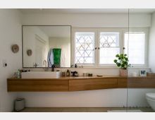 Modernes Badezimmer mit einem großen rechteckigen Spiegel über einem Holzwaschtisch. Auf dem Tisch steht ein weißes Waschbecken, Pflanzen, Pflegeprodukte und Dekorationsgegenstände; im Hintergrund sind drei Fenster mit Streben und halbtransparentem Sichtschutz zu sehen.