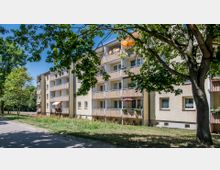 Mehrfamilienhaus mit beige gestrichener Fassade und mehreren Balkonen, die mit Markisen und Blumentöpfen dekoriert sind. Im Vordergrund befinden sich ein Gehweg, grüne Wiesen und mehrere Bäume, die Schatten spenden.