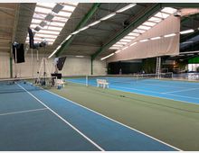 Tennis-Halle mit mehreren blau-grünen Plätzen, darüber ein gewölbtes Dach mit Oberlichtern. Beleuchtungsausrüstung, Leitern und Requisiten sind auf einem Platz aufgebaut, während weitere Ausrüstungsgegenstände und eine Bank im Hintergrund sichtbar sind.