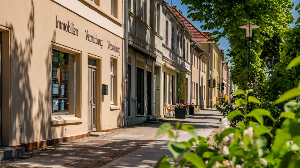 Straßenzug mit niedrigen, aneinandergebauten Altbaufassaden und Schaufenstern entlang eines breiten Gehwegs. Im Vordergrund rahmen grüne Bäume und Sträucher die Szene, daneben stehen Straßenlaterne und Ampel an der ruhigen Straße.