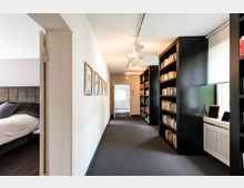 Ein moderner, heller Flur mit Teppichboden, schwarzen Bücherregalen an der rechten Wand und mehreren Büchern. Auf der linken Seite befindet sich eine geöffnete Tür, die in ein Schlafzimmer mit einem gepolsterten Bett führt. Im Hintergrund ist eine weitere Tür zu sehen, die zu einem anderen Raum führt.