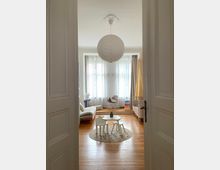 Kinderzimmer mit Holzboden und hellen Wänden, eingerichtet mit einem kleinen runden Tisch und passenden Stühlen auf einem Teppich. Im Hintergrund stehen ein Sofa, ein Kinderbett, ein Schaukelstuhl und ein Stapelturm vor bodentiefen Fenstern mit Vorhängen; eine große Papierlampe hängt von der Decke.