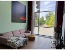 Wohnzimmer mit einer großen Fensterfront, die ins Grüne blickt. Der Raum enthält ein rosa Sofa, eine Decke mit Hirschmotiv, einen hölzernen Sitzpouf, Pflanzen in Töpfen, eine wandgroße Kunstmalerei und eine moderne Hängelampe mit Kugelleuchten.