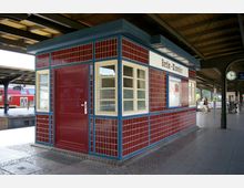 Bahnhofsgebäude am S-Bahnhof Berlin-Wannsee mit roter Kachelverkleidung, blauen Rahmen und weißen Fenstern; im Hintergrund ein roter Zug sowie eine Bahnhofsuhr unter einem Bahnsteigdach.