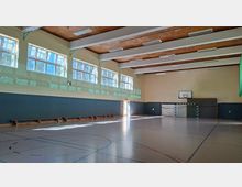 Große Sporthalle mit markiertem Hallenboden, langen Holzleistenbänken an der Wand und hohen Fensterreihen, die viel Tageslicht hereinlassen. An der Stirnseite sind ein Handballtor und ein Basketballkorb zu sehen, unter einer Decke mit Holzbalken und Deckenleuchten.