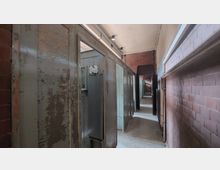 Ein schmaler Korridor in einem historischen Gebäude mit Zellen auf der linken Seite, die durch abgenutzte Holztüren mit kleinen Fenstern abgetrennt sind. Die Wände bestehen aus rotem Backstein, und die Decke ist schlicht verputzt mit vereinzelter Beleuchtung.