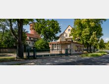Eingezäunter Eingangsbereich mit einer kleinen Wachhütte und grünen Toren, dahinter ein Wohngebäude mit Satteldach und ein roter Wasserturm. Umgeben von Bäumen und einer gepflasterten Straße im Vordergrund.
