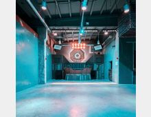 Leerer Clubraum mit industriellem Design, Betonböden und sichtbarer Deckenkonstruktion. Der Fokus liegt auf einer Bühne mit einem DJ-Pult aus Holzpanelen und einer geometrischen Wanddekoration, umgeben von Lautsprechern und moderner Beleuchtung.