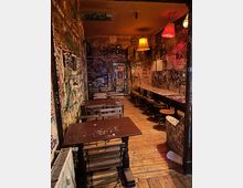 Schmaler Innenraum eines alternativ gestalteten Cafés oder Bars mit Holzdielenboden, langen Holztischen und Hockern entlang der rechten Wand sowie Bänken links. Die Wände und die Tür sind dicht mit Graffiti, Stickern und Zeichnungen bedeckt, beleuchtet von mehreren hängenden Lampenschirmen in warmen Farben.