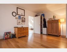 Ein schlicht eingerichtetes Zimmer mit Holzboden, einer Kommode und einem Schrank aus Holz. An der weißen Wand hängen diverse Bilderrahmen, und durch eine offene Tür ist ein angrenzender Raum mit Dachfenstern zu sehen.