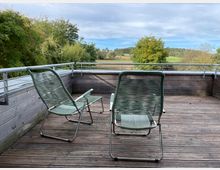 Zwei grüne Gartenstühle aus Metall und Kunststoffgeflecht stehen auf einer Terrasse mit Holzdielen, die von einer hölzernen Brüstung mit Metallgeländer umrandet ist. Im Hintergrund befinden sich grüne Bäume und eine weite, ländliche Landschaft unter bewölktem Himmel.