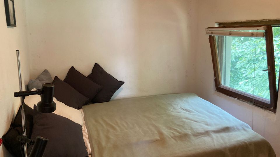 Kleines Schlafzimmer mit Doppelbett, dunklen Kissen und grünlicher Decke, flankiert von einer Stehlampe. Rechts ein Holzfenster mit teilweiser Jalousie und Blick auf dichtes grünes Laub draußen, dazu helle Wände und Holzdecke.