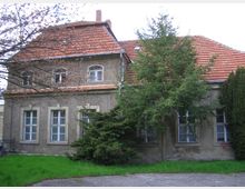 Zweistöckiges historisches Wohngebäude mit roten Ziegeldächern, rechteckigen Fenstern und grob verputzter Fassade, umgeben von grünen Bäumen und Rasen. Im Vordergrund sind eine gepflasterte Fläche und mehrere Nadelbäume sichtbar.