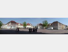 Breiter Platz in einer historischen Altstadt mit Kopfsteinpflaster, flankiert von klassizistischen Gebäuden mit roten Dächern und weißer/sandfarbener Fassade. Im Zentrum befinden sich Bäume, Cafétische und eine Gruppe von Menschen, die das sonnige Wetter genießen.