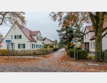 Wohnsiedlung mit gepflasterter Straße, von Laub bedeckt, gesäumt von Einfamilienhäusern mit roten Ziegeldächern und grünen Fensterläden. Im Vordergrund stehen Straßenschilder und ein großer Baum, der den Gehweg flankiert.