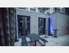 Innenhof-Terrasse eines modernen Gebäudes mit hellen Betonwänden und großen Glas-Schiebetüren. Im Vordergrund stehen ein schwarzer Picknicktisch mit Bänken und zwei Stehtische; hohe Kakteen und blaues Akzentlicht markieren die Ecken.
