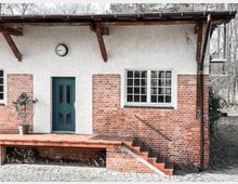 Außenansicht eines kleinen Gebäudes mit einer gemauerten Fassade aus roten Ziegelsteinen und verputzten weißen Flächen. Es gibt eine grüne Tür, ein großes Fenster mit Sprossen und eine kleine Treppe, die zu einem erhöhten Eingangsbereich mit Fliesen führt. Ein Holzdach mit sichtbaren Balken und eine Uhr über der Tür sind ebenfalls sichtbar; unter dem Eingangsboden lagert Brennholz.