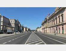 Breite Straße in einer urbanen Umgebung mit historischen Gebäuden auf beiden Seiten, darunter ein großes rosa-farbenes Gebäude mit Skulpturen an der Fassade. Im Hintergrund führt die Straße auf eine Baumreihe zu, bei klarem Himmel und wenig Verkehr.
