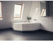 Modernes Badezimmer mit weißen, glänzenden Fliesen an den Wänden und grauem Fliesenboden. Eine Eckbadewanne mit schrägen Dachfenstern darüber, durch die Tageslicht einfällt; auf der Wanne steht eine Vase mit grünen Zweigen.