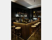 Eine gemütliche Bar mit dunklen Wänden, dezenter Beleuchtung und einer Theke mit vertikalen Holzleisten. Auf der Theke stehen Blumen, Gläser und Kerzen; im Hintergrund sind Regale mit Flaschen und Gläsern sowie hohe Hocker um die Theke verteilt.