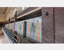 Innenansicht eines historischen Schwimmbades mit alten Umkleidekabinen aus Holz, deren Farbe abgeblättert ist. Die Backsteinwände, ein schmiedeeisernes Geländer und buntglasartige Fenster oberhalb der Kabinen verleihen dem Raum eine nostalgische Atmosphäre.