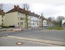 Straßenszene mit mehrstöckigen Wohnhäusern im Hintergrund, die Reihenhäuser mit hellen Fassaden und roten Ziegeldächern zeigen. Im Vordergrund ist eine Baustelle mit Absperrungen und Warnschildern, daneben eine schmale Grünfläche mit kahlen Bäumen entlang der Straße.