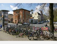 Straßenszene in einer urbanen Umgebung mit einem roten Backsteingebäude im Hintergrund. Mehrere Fahrräder stehen dicht an einem Fahrradständer, während Autos und Personen die Straße überqueren; links ist eine Litfaßsäule zu sehen.