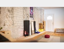 Innenraum mit freigelegter, teils verputzter Ziegelwand und Holzboden, eingerichtet mit Teppich, rotem Sitzkissen, Standspiegel, zwei hohen Lautsprechern und gestapelten Holzstühlen. Rechts befindet sich eine weiße Tür mit Fenster sowie ein niedriges Schuhregal.