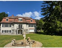 Zweistöckiges Haus im klassischen Stil mit hellen Fassaden, Schieferdach und zentralem Balkon über einem Eingangsbogen aus Stein. Im Vordergrund befindet sich ein Brunnen mit einer Statue, umgeben von einer Rasenfläche und großen Bäumen.
