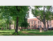 Roter Backsteinbau mit großen Rundbogenfenstern im Hintergrund, umgeben von einer Grünfläche mit hohen Bäumen und Sitzgelegenheiten. Der Garten hat einen gepflasterten Weg und vereinzelt sichtbare Backsteinsäulen.