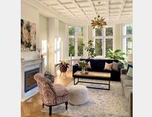 Großes, helles Wohnzimmer mit einem eleganten Marmorkamin, einer Sitzgruppe aus einem blauen Sofa, einem gemusterten Sessel und einem Holztisch. Das Zimmer hat ein kassettengemustertes Deckendesign, große Fenster, helle Wände und Pflanzen, die das natürliche Licht betonen.