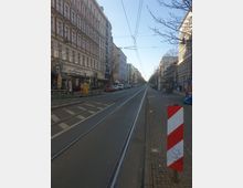 Straßenansicht mit Schienen einer Straßenbahn, flankiert von mehrstöckigen Wohn- und Geschäftshäusern im Altbaustil. Im Vordergrund steht ein rot-weißes Warnschild, und die Straße ist von parkenden Autos und kahlen Bäumen gesäumt.