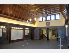 Eingangsbereich eines historischen Bahnhofs mit dekorativer Holzdecke, blauen Wandfliesen und großen Anzeigetafeln. Im Hintergrund befinden sich geöffnete Holztüren, die zu einem Treppenabgang führen, beleuchtet von klassischen Decken- und Wandlampen.