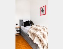 Ein schlichtes Schlafzimmer mit einem grauen Polsterbett, weißer Bettwäsche, schwarzen Kissen und einer braunen Kunstfelldecke. An der weißen Wand hängt ein gerahmtes Bild mit Hahnmotiv in einem roten Rahmen, daneben ein kleiner Nachttisch mit einer Lampe.