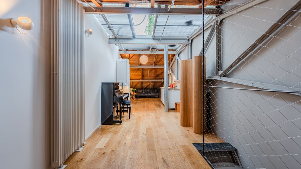 Langer, schmaler Innenraum im Loft-Stil mit hellem Holzboden, weiß gestrichenen Wänden und sichtbarer Holzbalkendecke unter einem Oberlicht. Rechts begrenzt ein Drahtnetz eine Treppe, links stehen ein hoher Heizkörper und ein Klavier; im Hintergrund sind eine kleine Küchenzeile und ein Sitzbereich zu sehen.