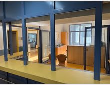 Blick durch ein Fenster mit blauer Metallrahmung und gelber Fensterbank in ein modernes Büro mit Holzmöbeln. Im Hintergrund sind eine Glastür, ein Schreibtisch, ein Schrank und helle Wände sichtbar, durchzogen von natürlichem Licht.