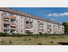 Mehrstöckiges Wohngebäude in schlichtem Putzbau mit mehreren Balkonen, von denen einige Markisen oder Pflanzen haben. Im Vordergrund befindet sich eine Wiese mit vereinzelten Sträuchern, der Himmel ist blau mit ein paar Wolken.