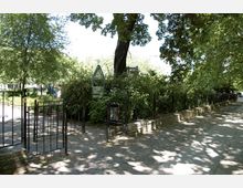 Ein kleiner Parkeingang mit schwarzen Metallgittern, umgeben von dichtem Grün. Ein Weg aus Pflastersteinen führt entlang einer Baumreihe, die Schatten auf den Boden wirft.