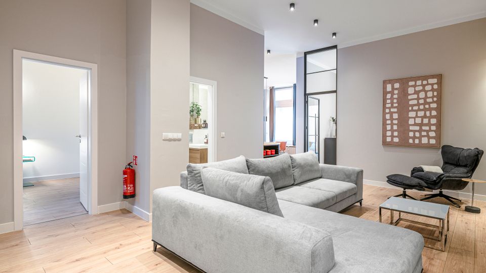 Modernes, offenes Wohnzimmer mit hellgrauem Ecksofa auf Holzfußboden, Glas-Couchtisch und schwarzem Relaxsessel neben einem Wandbild. Hohe Decken mit Einbauspots, breite Türöffnungen und eine Glastrennwand führen zu angrenzenden Räumen und einem hellen Fensterbereich.