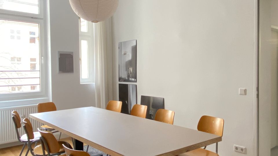 Heller Besprechungs- oder Essraum mit großem rechteckigem Tisch auf Teppich und mehreren Holzstühlen mit Metallgestell. Hohe Fenster mit Vorhängen, weiße Wände, eine runde Papier-Hängelampe und ein Türdurchgang in einen angrenzenden Raum sind sichtbar.