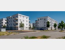 Mehrfamilienhäuser in moderner Bauweise mit weißen Fassaden und Balkonen, gelegen in einer sauberen Wohngegend mit gepflasterten Straßen, vereinzelt geparkten Autos und kleinen Bäumen entlang der Gehwege. Der Himmel ist klar und blau.