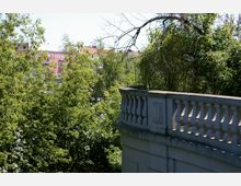 Eine steinerne Balustrade in einem parkähnlichen Bereich, umgeben von üppigem Laubwerk und Bäumen; im Hintergrund sind farbige Wohngebäude mit roten Dächern sichtbar.